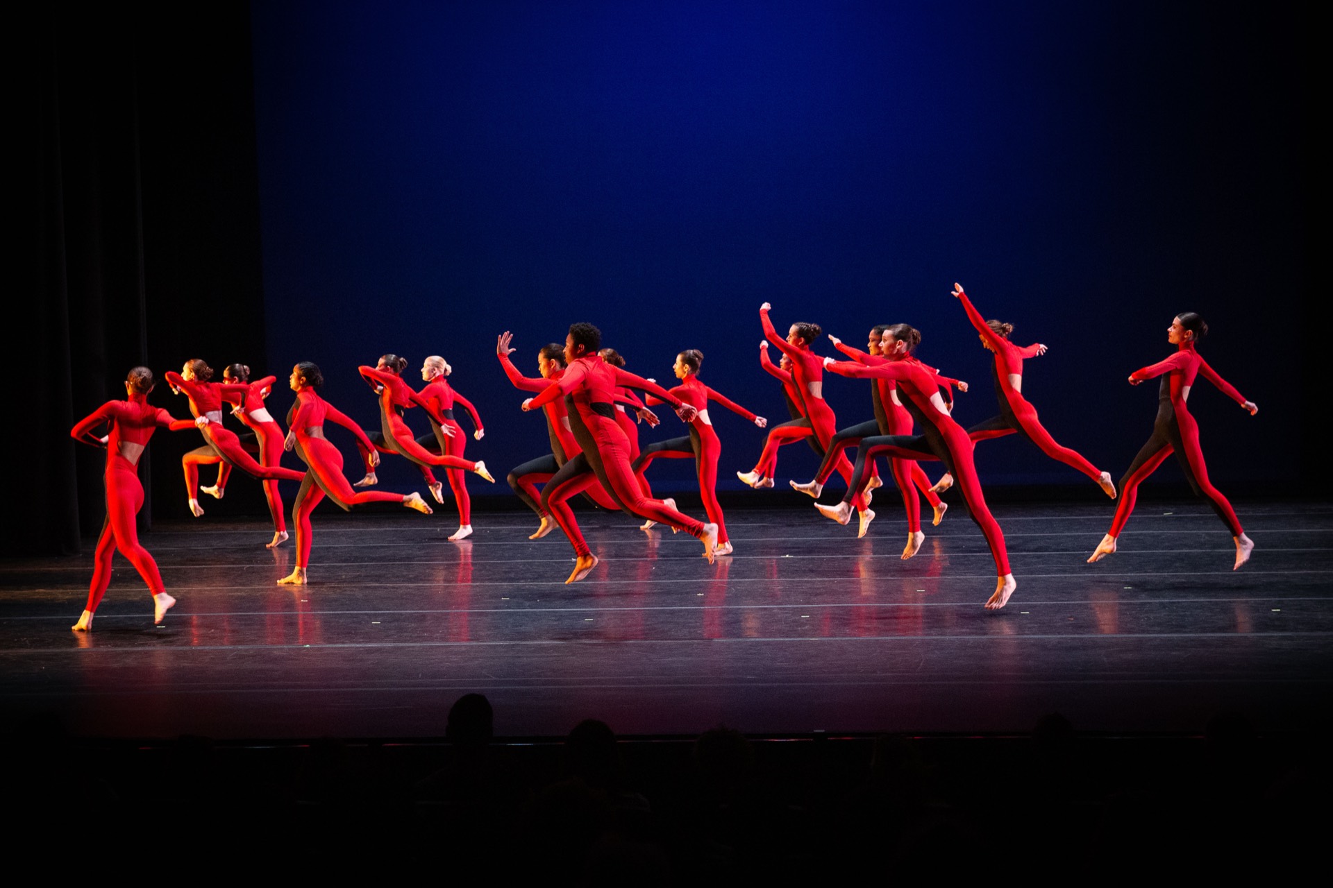 TWDCC dancers in red leap together across a dark stage.